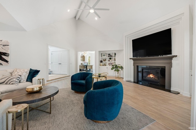 living area featuring high vaulted ceiling, a glass covered fireplace, a ceiling fan, wood finished floors, and beamed ceiling