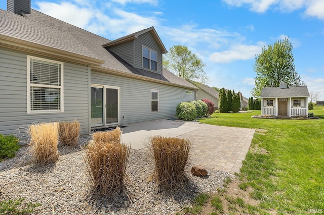 rear view of property featuring an outdoor structure, a patio area, roof with shingles, and a yard