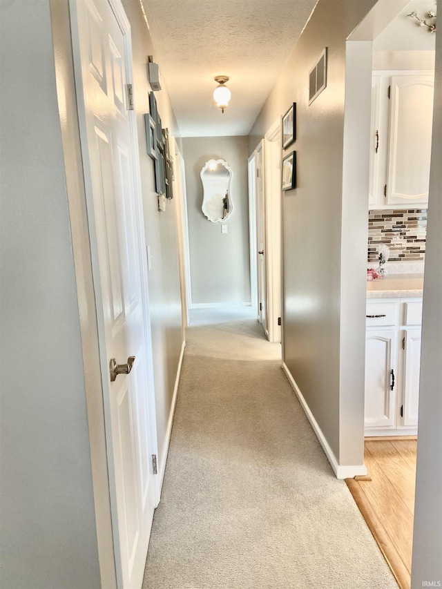 hallway with visible vents, a textured ceiling, baseboards, and light carpet