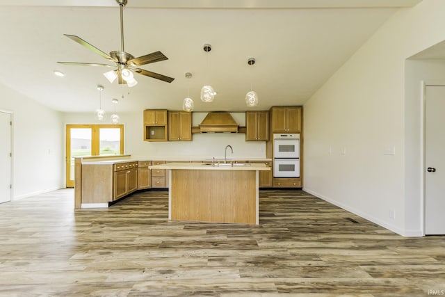 kitchen with a center island with sink, wood finished floors, premium range hood, and white double oven