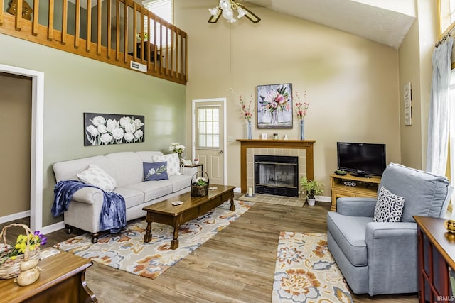 living room featuring ceiling fan, a tiled fireplace, wood finished floors, baseboards, and high vaulted ceiling