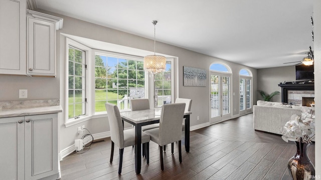 dining room with baseboards, ceiling fan with notable chandelier, wood finished floors, and a warm lit fireplace