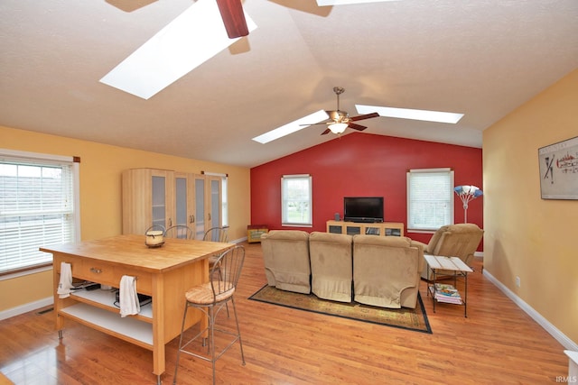 living room with baseboards, a ceiling fan, light wood-style flooring, and vaulted ceiling with skylight