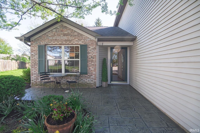 doorway to property featuring brick siding, a shingled roof, fence, and a patio