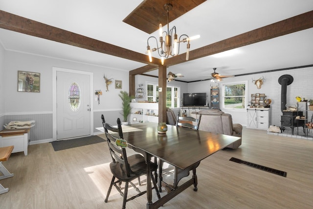 dining space with visible vents, a wood stove, beam ceiling, light wood-style flooring, and ceiling fan with notable chandelier