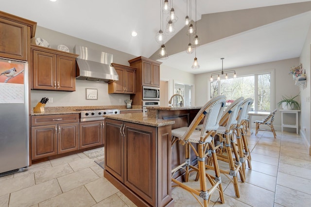 kitchen featuring light stone countertops, a breakfast bar, a center island with sink, wall chimney range hood, and stainless steel appliances