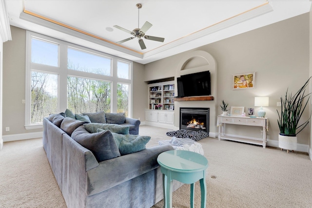 carpeted living area with a ceiling fan, a lit fireplace, a raised ceiling, baseboards, and ornamental molding