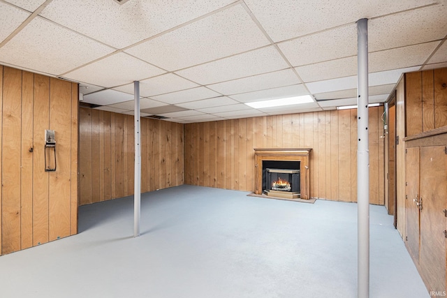 finished below grade area featuring wood walls, a drop ceiling, and a warm lit fireplace