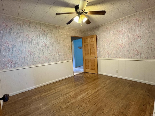 empty room with a ceiling fan, wallpapered walls, wainscoting, and wood finished floors