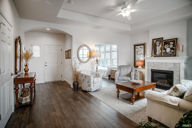 living area featuring a ceiling fan, arched walkways, a fireplace, wood finished floors, and a tray ceiling