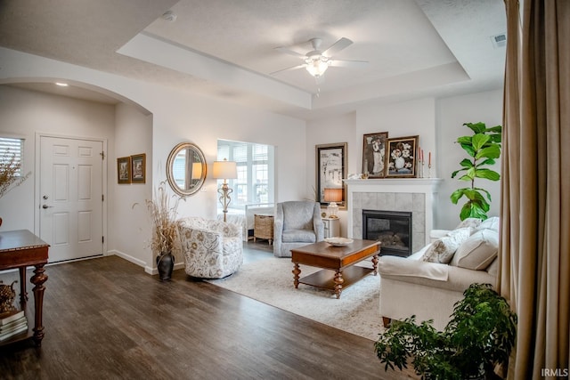 living area featuring a ceiling fan, a tile fireplace, a raised ceiling, baseboards, and dark wood-style floors
