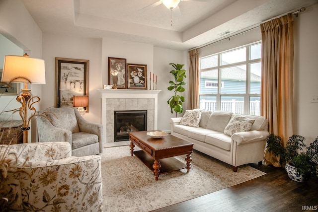 living room featuring a raised ceiling, a tiled fireplace, a ceiling fan, wood finished floors, and visible vents