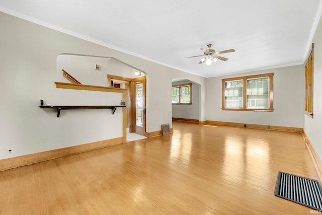 unfurnished living room featuring a ceiling fan, crown molding, arched walkways, wood finished floors, and baseboards