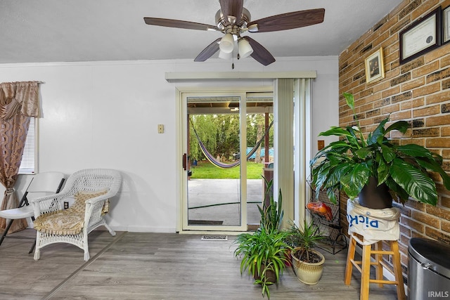 interior space featuring ceiling fan, brick wall, baseboards, wood finished floors, and ornamental molding