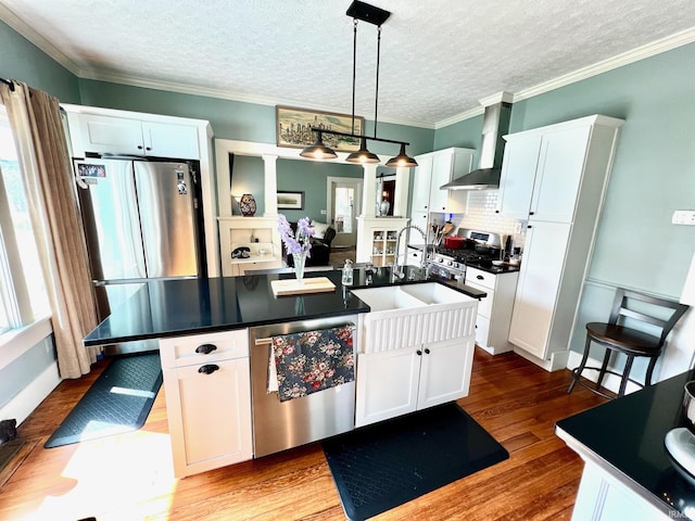 kitchen featuring wall chimney range hood, white cabinets, dark countertops, an island with sink, and stainless steel appliances
