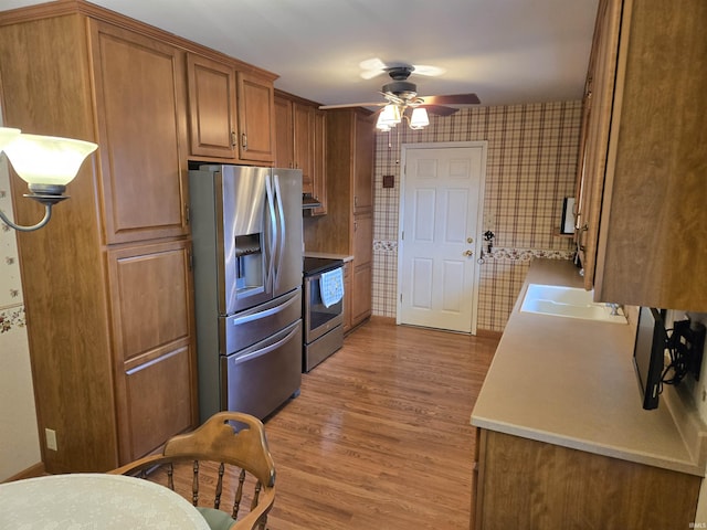 kitchen featuring appliances with stainless steel finishes, brown cabinetry, light wood-style flooring, a ceiling fan, and wallpapered walls