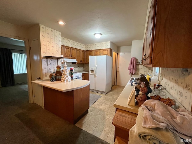 kitchen with brown cabinetry, a peninsula, under cabinet range hood, wallpapered walls, and white appliances