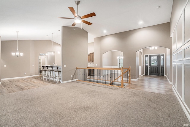 unfurnished living room featuring light colored carpet, arched walkways, high vaulted ceiling, a ceiling fan, and baseboards