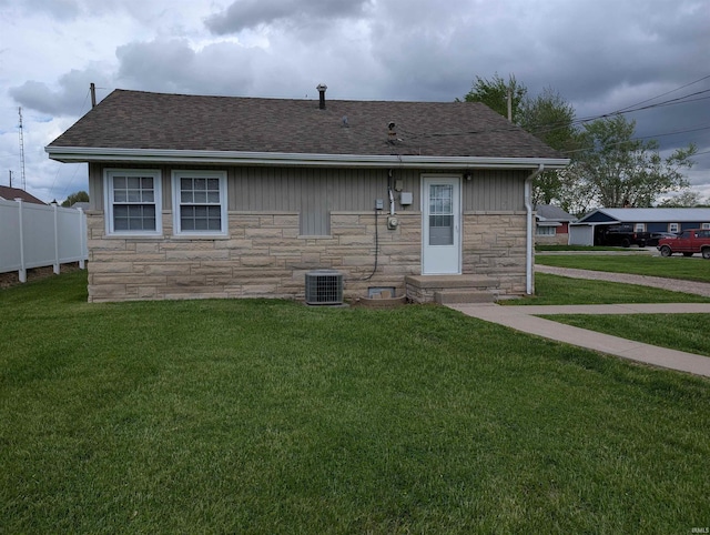 rear view of property with fence, stone siding, cooling unit, roof with shingles, and a yard