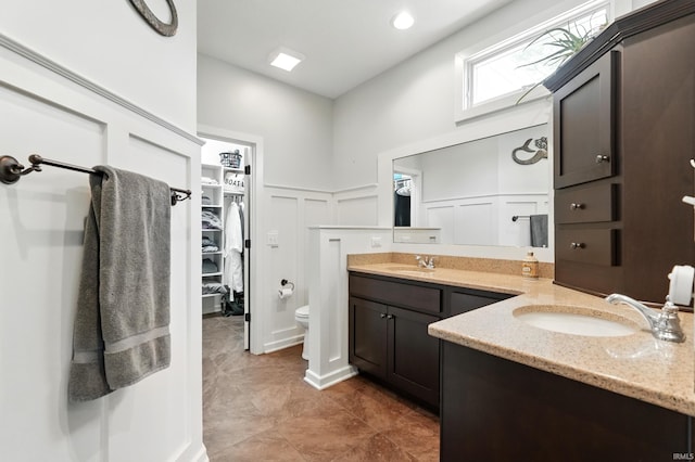full bathroom featuring a decorative wall, double vanity, wainscoting, a spacious closet, and toilet