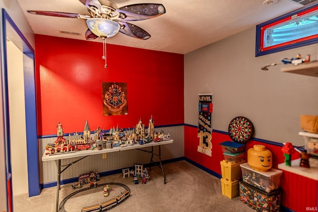 game room with carpet, a wainscoted wall, a textured ceiling, visible vents, and ceiling fan