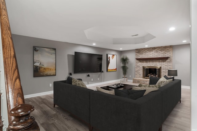 living room featuring wood finished floors, recessed lighting, a brick fireplace, a tray ceiling, and baseboards