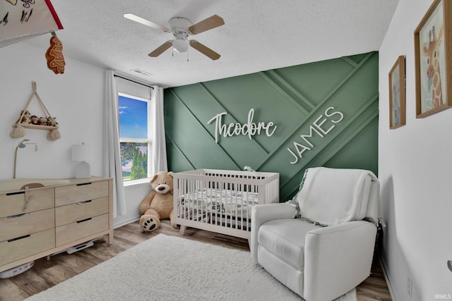 bedroom with a textured ceiling, a nursery area, visible vents, ceiling fan, and wood finished floors