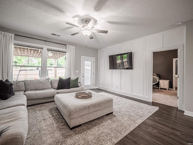 living room featuring a decorative wall, a textured ceiling, visible vents, dark wood-style flooring, and ceiling fan