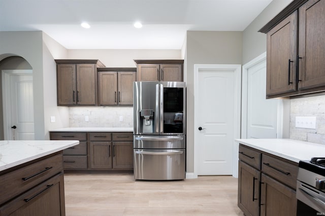 kitchen with stainless steel refrigerator with ice dispenser, light stone countertops, arched walkways, light wood-style floors, and tasteful backsplash
