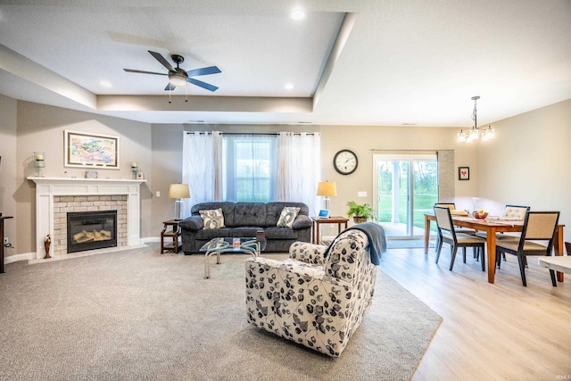 living room with a brick fireplace, a tray ceiling, light wood-style flooring, and ceiling fan with notable chandelier