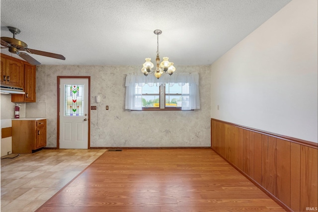 unfurnished dining area featuring light wood-style flooring, a chandelier, a ceiling fan, a textured ceiling, and wainscoting