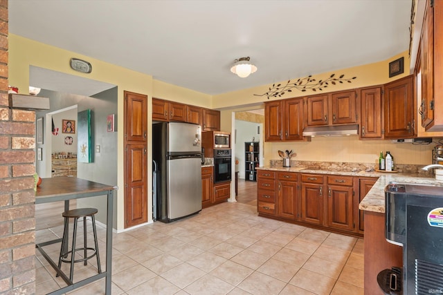kitchen featuring appliances with stainless steel finishes, under cabinet range hood, light tile patterned floors, brown cabinets, and light stone countertops