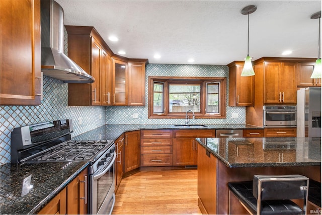 kitchen featuring appliances with stainless steel finishes, a sink, wall chimney range hood, brown cabinets, and light wood-type flooring