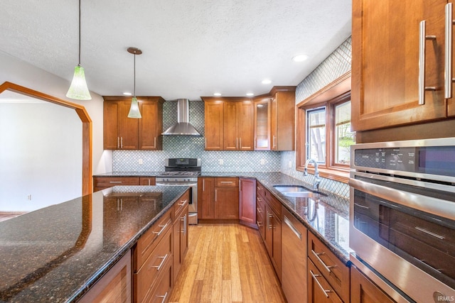 kitchen with wall chimney exhaust hood, a sink, appliances with stainless steel finishes, brown cabinetry, and light wood-style floors