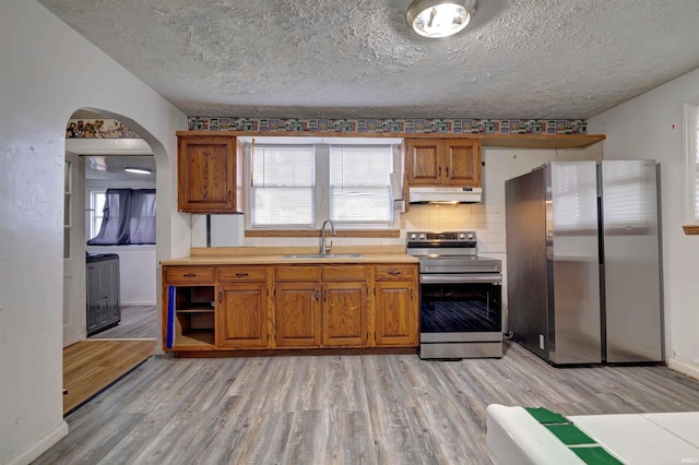 kitchen with appliances with stainless steel finishes, a sink, under cabinet range hood, light wood-style flooring, and brown cabinets