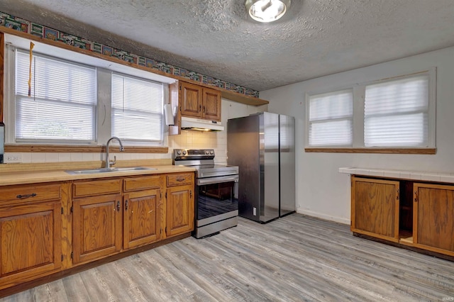 kitchen featuring stainless steel appliances, a sink, under cabinet range hood, brown cabinets, and light wood-type flooring