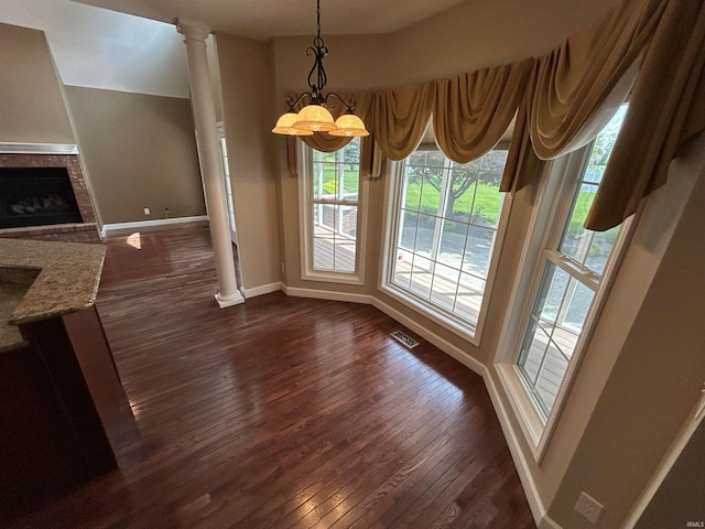unfurnished dining area featuring baseboards, dark wood-style flooring, decorative columns, a chandelier, and a fireplace