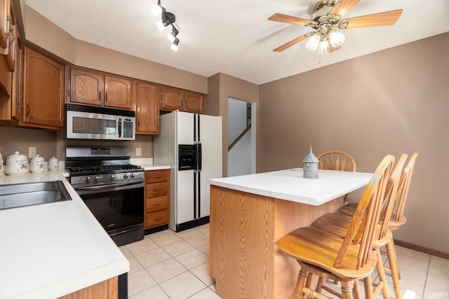 kitchen with stainless steel appliances, light tile patterned floors, light countertops, brown cabinetry, and ceiling fan