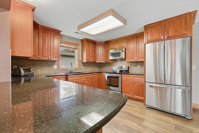 kitchen with appliances with stainless steel finishes, a sink, light wood finished floors, tasteful backsplash, and dark stone countertops