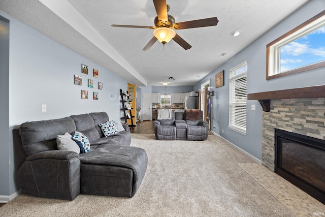 living room with a stone fireplace, a ceiling fan, light carpet, baseboards, and a textured ceiling