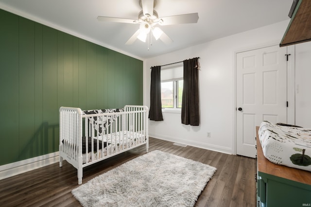 bedroom featuring a nursery area, dark wood-style floors, baseboards, and ceiling fan