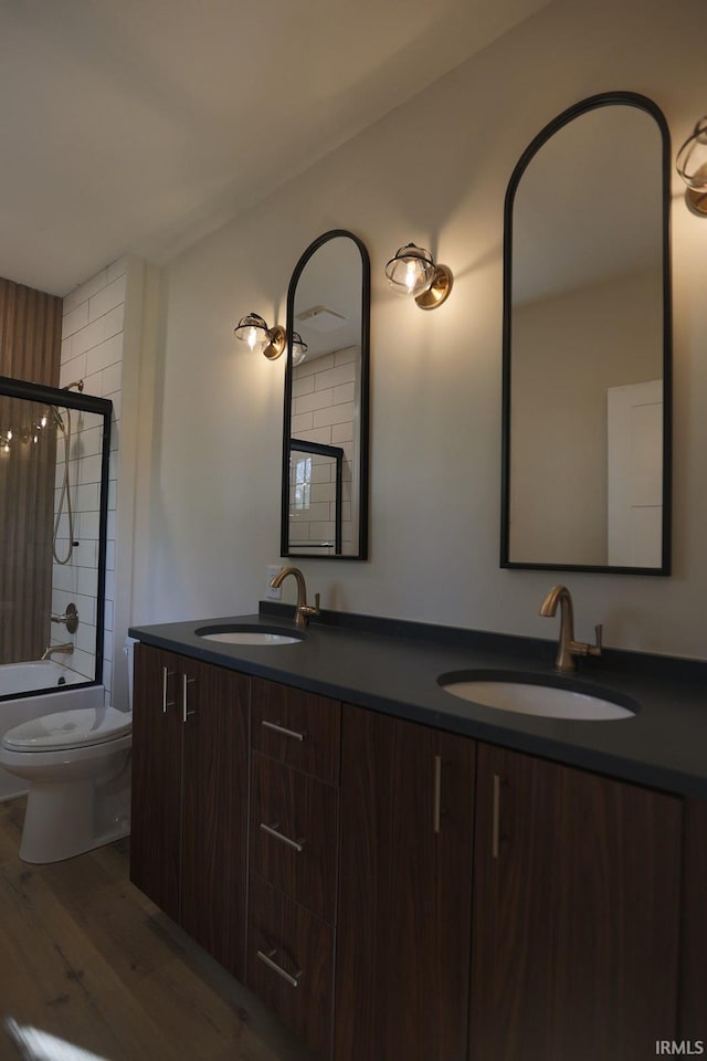 bathroom featuring wood finished floors, double vanity, toilet, and bath / shower combo with glass door