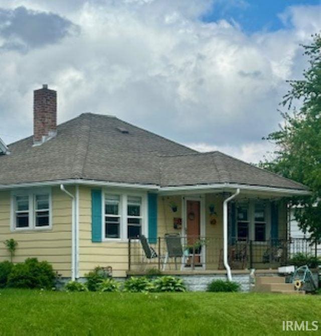 view of front of house with a chimney, covered porch, a front lawn, and a shingled roof