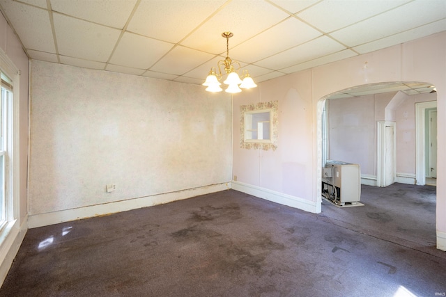 unfurnished dining area with arched walkways, a drop ceiling, dark colored carpet, baseboards, and a chandelier