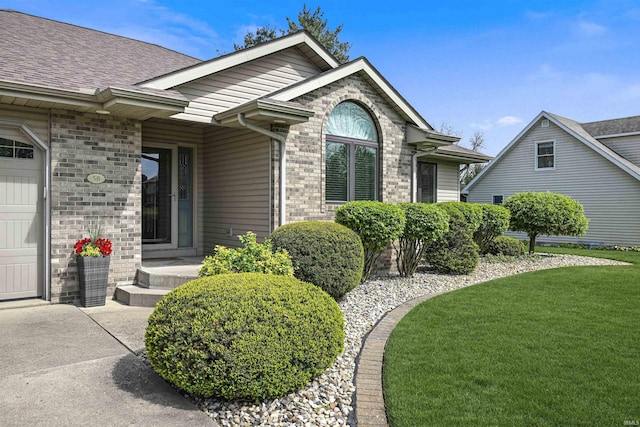 view of exterior entry with a yard, brick siding, a shingled roof, driveway, and an attached garage