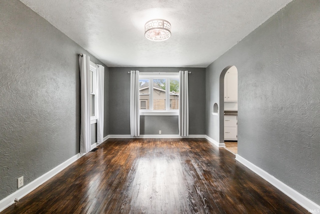 empty room featuring a textured wall, arched walkways, wood finished floors, a textured ceiling, and baseboards