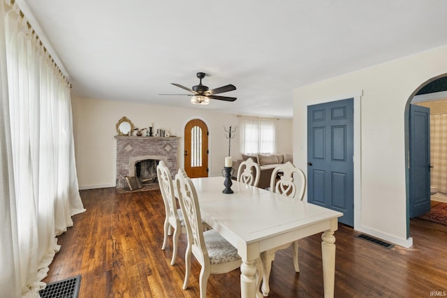 dining room featuring arched walkways, a ceiling fan, a brick fireplace, dark wood finished floors, and baseboards