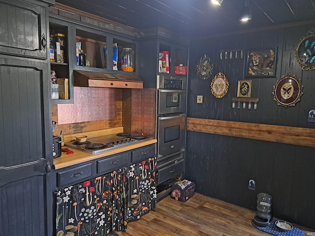 kitchen with stainless steel electric stovetop, double oven, under cabinet range hood, wood finished floors, and wood walls