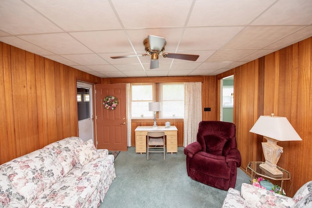 carpeted living area with wood walls, ceiling fan, and a paneled ceiling
