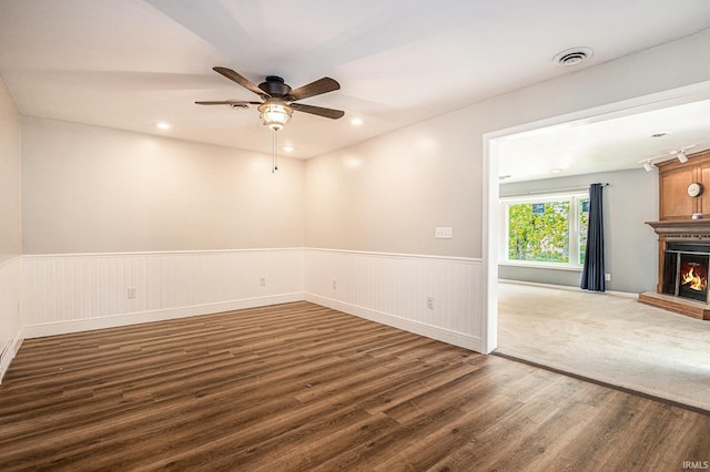 unfurnished room featuring a warm lit fireplace, dark wood finished floors, a wainscoted wall, a ceiling fan, and recessed lighting